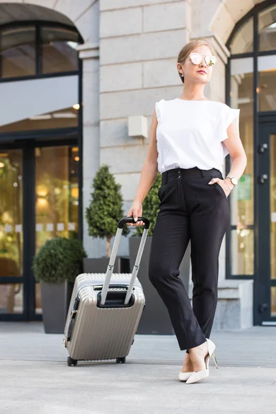 Una chica en un traje de negocios y gafas con una maleta en el fondo de un hotel caro. joven hermosa mujer eset equipaje — Foto de Stock