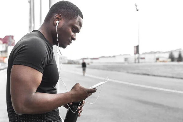Jeune athlète de jogging masculin s'entraînant et faisant de l'entraînement à l'extérieur en ville. un homme noir se reposant après une séance d'entraînement, écoutant de la musique et regardant une montre de sport — Photo
