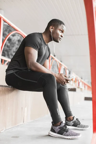 Young male jogger athlete training and doing workout outdoors in city. a black man resting after a workout and listening to music and watching a sports watch