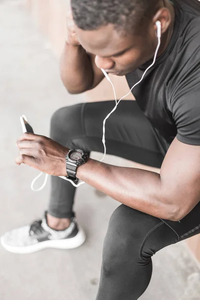 Young male jogger athlete training and doing workout outdoors in city. a black man resting after a workout and listening to music and watching a sports watch