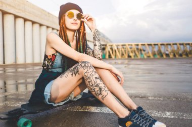 dövme ve dreadlocks mavi şapkalı bir genç kızla bir longboard beton yapı çerçevede oturur