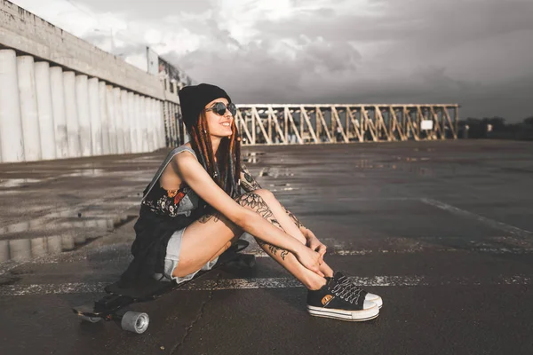 Jovem com tatuagens e dreadlocks em um boné azul senta-se em um longboard contra o fundo da estrutura de concreto — Fotografia de Stock
