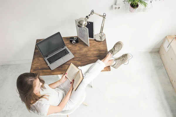 Νεαρή γυναίκα εργασία στο σπίτι και διαβάζοντας ένα βιβλίο με τα πόδια της πάνω στο τραπέζι. Δημιουργική σκανδιναβικό στυλ χώρου εργασίας. Wiew από ψηλά — Φωτογραφία Αρχείου