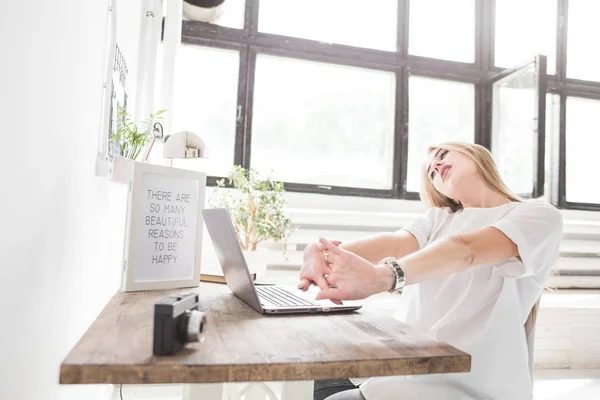 Νεαρή γυναίκα εργάζεται στο σπίτι πίσω από ένα φορητό υπολογιστή και τεντώνοντας τα χέρια της. Δημιουργική σκανδιναβικό στυλ χώρου εργασίας — Φωτογραφία Αρχείου
