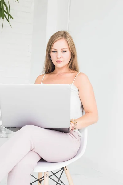 Портрет молодої блондинки, що сидить на стільці на білій стіні і працює в ноутбуці — стокове фото