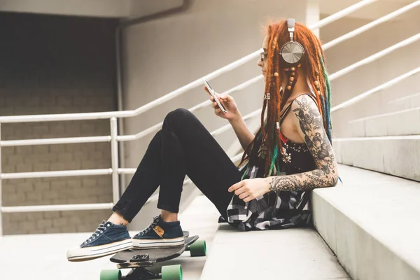 Chica joven con tatuaje y rastas escuchando música mientras se sienta en los escalones —  Fotos de Stock