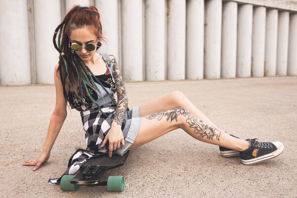 young girl with tattoo and dreadlocks sitting on a longboard on the background of concrete structure