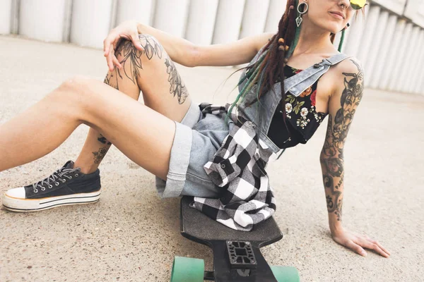 Chica joven con tatuaje y rastas sentado en un longboard en el fondo de la estructura de hormigón —  Fotos de Stock