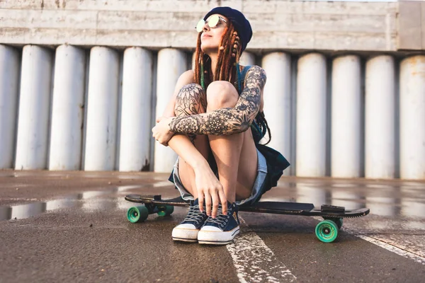 Young girl with tattoos and dreadlocks in a blue cap sits on a longboard against the background of concrete structure — Stock Photo, Image