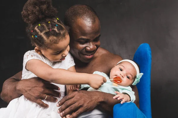 Musculoso hombre afroamericano sentado con niños en una silla azul contra una pared negra — Foto de Stock