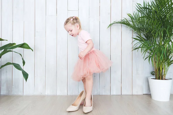 Linda niña con sus zapatos de madres mientras está de pie junto a las plantas en macetas sobre un fondo blanco . — Foto de Stock