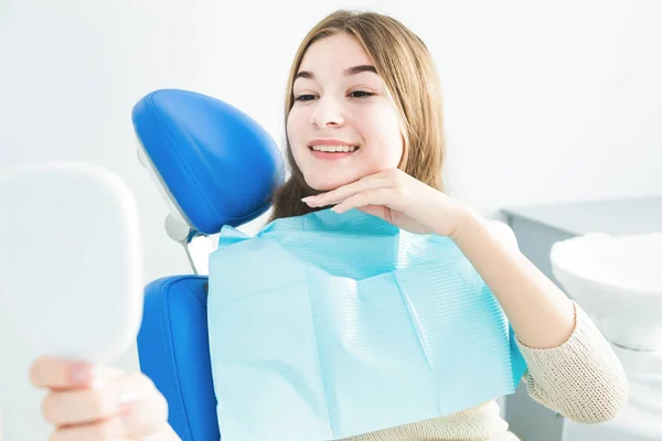 Clínica dentária. Recepção, exame do paciente. Cuidado com os dentes. Menina sorrindo, olhando no espelho depois de um check-up dental em seu dentista — Fotografia de Stock