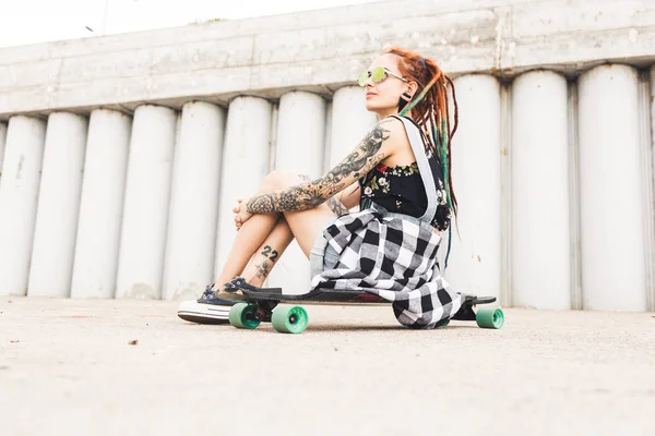 Young girl with tattoo and dreadlocks sitting on a longboard on the background of concrete structure — Stock Photo, Image