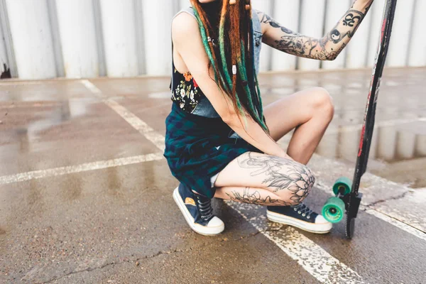 Jovem com tatuagens e dreadlocks senta-se com um longboard no fundo da estrutura de concreto — Fotografia de Stock