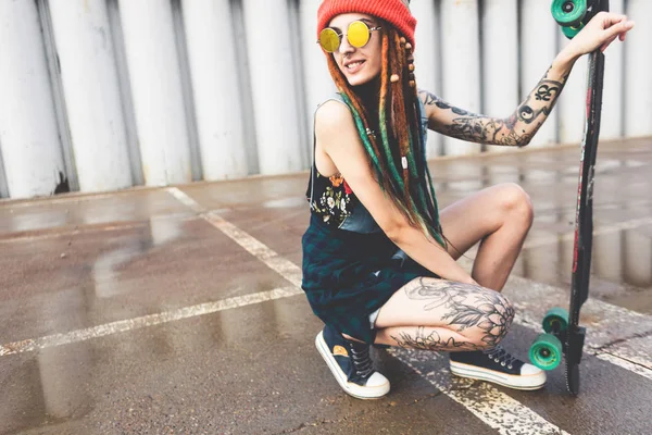 Young girl with tattoos and dreadlocks in a cap sits with a longboard on the background of concrete structure — Stock Photo, Image