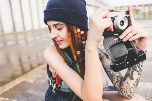 Молодая девушка с татуировками и дредами в синей шляпе фотографирует винтажную камеру на фоне бетонной стены — стоковое фото