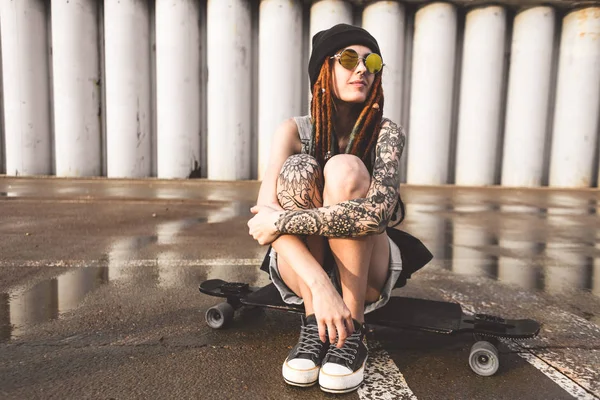 Young girl with tattoos and dreadlocks in a blue cap sits on a longboard against the background of concrete structure — Stock Photo, Image