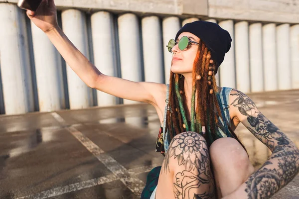 Young girl with tattoos and dreadlocks in a blue cap uses a smartphone against the background of a concrete wall — Stock Photo, Image