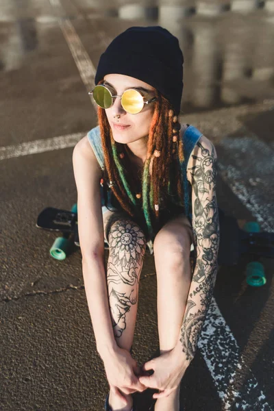 Young girl with tattoos and dreadlocks in a blue cap sits on a longboard against the background of concrete structure — Stock Photo, Image