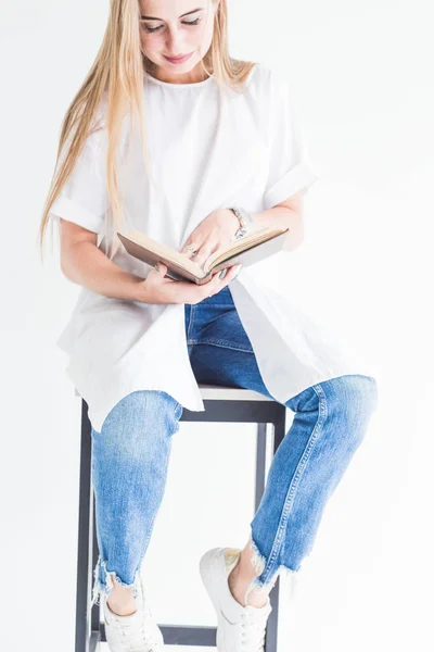 Retrato de una joven rubia con estilo en una camiseta blanca y jeans azules leyendo un libro sobre un fondo blanco — Foto de Stock