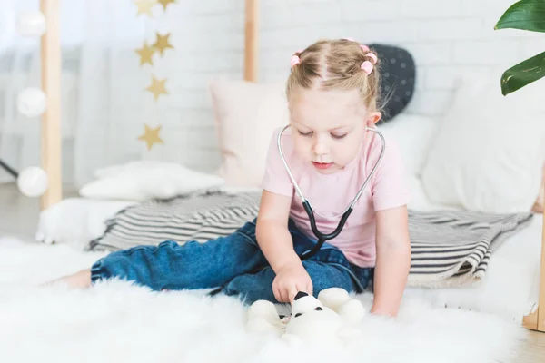 Linda niña sentada en la cama y jugando al doctor con estetoscopio y oso de peluche . — Foto de Stock