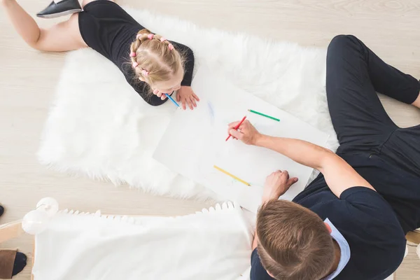 Un joven padre y su hija yacen en el suelo y dibujan con lápices de colores . — Foto de Stock