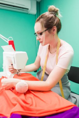 Dikiş atölyesi. Terzi iş başında. Dikiş makinesi ile renkli bir arka plan üzerine çalışan genç kadın