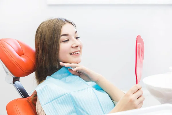 Clínica dental. Recepción, examen del paciente. Cuidado de los dientes. Chica joven sonriendo, mirando en el espejo después de un chequeo dental a su dentista — Foto de Stock