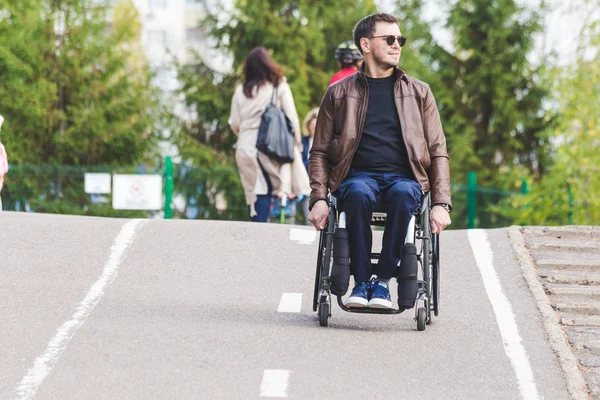 Un giovane su una sedia a rotelle cavalca lungo la strada del parco . — Foto Stock