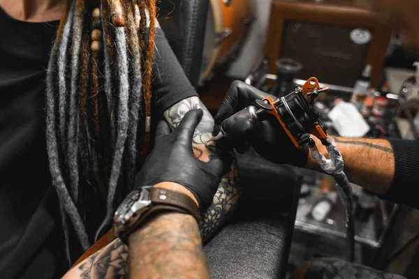 Fille avec dreadlocks dans un salon de tatouage. Le maître crée une image sur le corps d'une jeune belle fille. Gros plan des mains et de la machine à tatouer — Photo