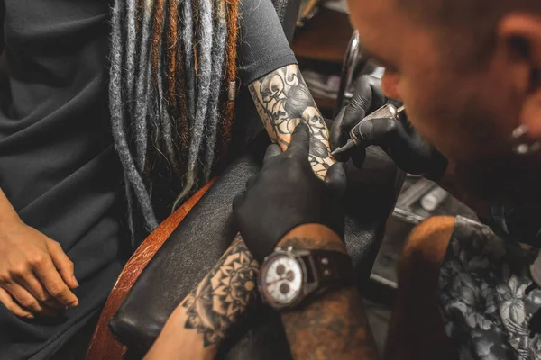 Fille avec dreadlocks dans un salon de tatouage. Le maître crée une image sur le corps d'une jeune belle fille. Gros plan des mains et de la machine à tatouer — Photo