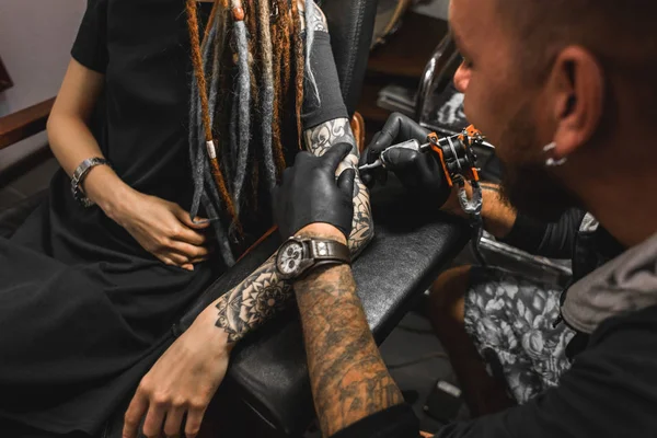 Fille avec dreadlocks dans un salon de tatouage. Le maître crée une image sur le corps d'une jeune belle fille. Gros plan des mains et de la machine à tatouer — Photo