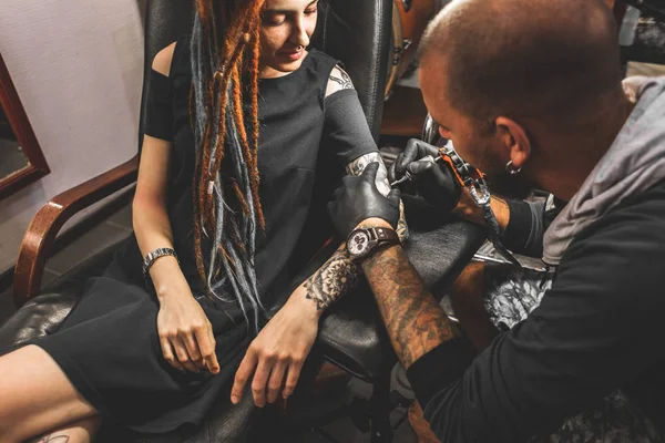 Lány a raszta a egy tetováló szalonnal. A mester egy képet hoz létre, a test egy fiatal gyönyörű lány. Közeli kép a kéz és a tetováló gép — Stock Fotó