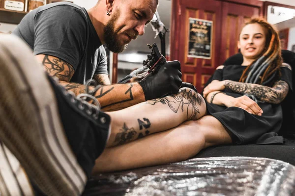 Menina com dreadlocks em uma sala de tatuagem. O mestre cria uma imagem no corpo de uma jovem menina bonita. Close-up de mãos e máquina de tatuagem — Fotografia de Stock