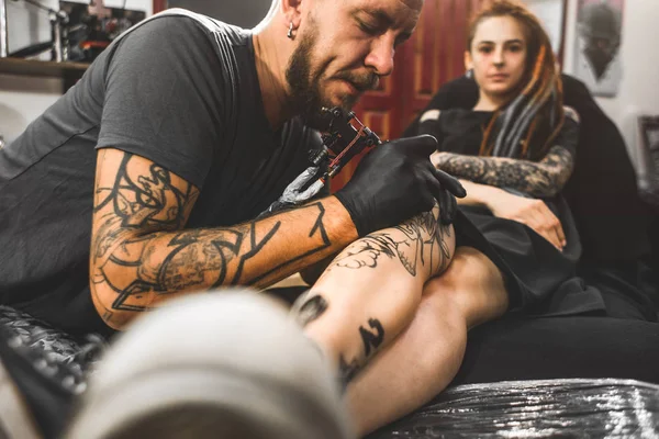 Fille avec dreadlocks dans un salon de tatouage. Le maître crée une image sur le corps d'une jeune belle fille. Gros plan des mains et de la machine à tatouer — Photo