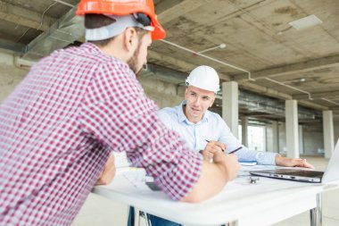 Erkek mühendisler, mimarlar kask masasında çalışıyor. Çizimler, dizüstü bilgisayar, masaüstü rulet. Resepsiyon ve inşaat yapı denetim