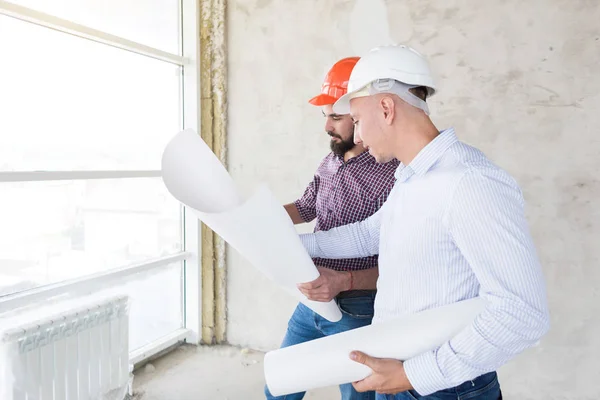 Ingenieros masculinos, arquitectos en cascos se paran junto a la ventana y sostienen un rollo de papel con un dibujo del edificio — Foto de Stock