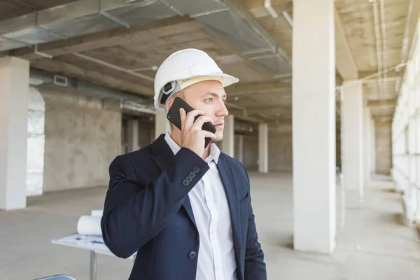 male engineer, foreman in a helmet, in a business suit with paper with drawings, in the construction being completed talking on mobile phone. building supervision