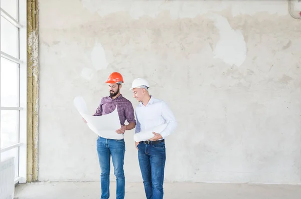 Ingenieros masculinos, arquitectos en cascos se paran junto a la ventana y sostienen un rollo de papel con un dibujo del edificio — Foto de Stock