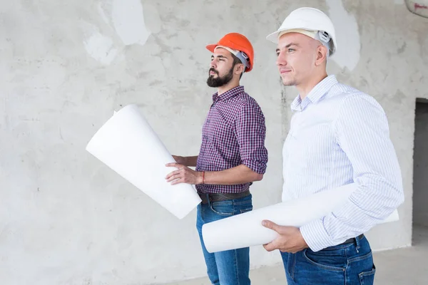 Ingenieros masculinos, arquitectos en cascos se paran junto a la ventana y sostienen un rollo de papel con un dibujo del edificio — Foto de Stock