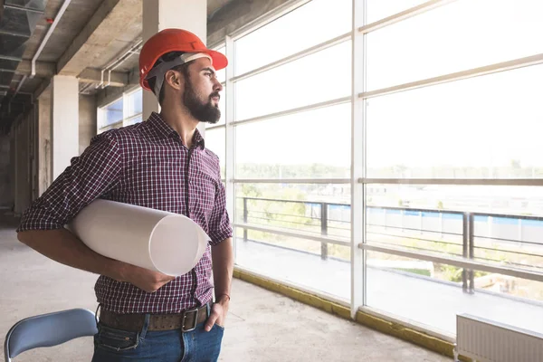 El hombre es un ingeniero en un casco, en una camisa a cuadros con papel con dibujos, en la construcción que se está completando. supervisión de edificios — Foto de Stock