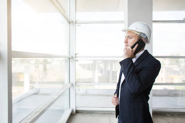 Ingeniero masculino, capataz en un casco, en un traje de negocios con papel con dibujos, en la construcción que se completa hablando por teléfono móvil. supervisión de edificios — Foto de Stock