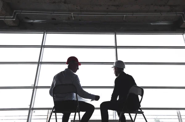 Hombres ingenieros constructores que trabajan en la mesa con un ordenador portátil, negociaciones comerciales en el sitio de construcción. siluetas contra el fondo de la ventana. ganga, compra de apartamento, bienes raíces — Foto de Stock