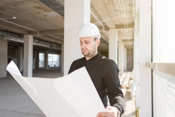 El arquitecto constructor en un casco con dibujos está trabajando en el sitio de construcción. supervisión de la construcción . — Foto de Stock