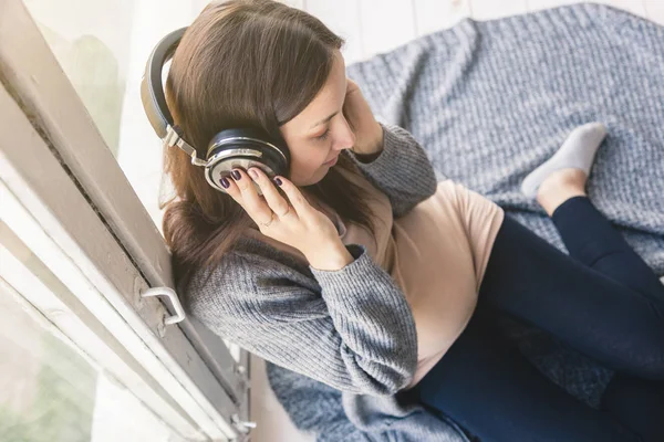 Mujer embarazada joven escuchando música y sentada en el sofá de la habitación. música clásica para el bebé en el útero — Foto de Stock