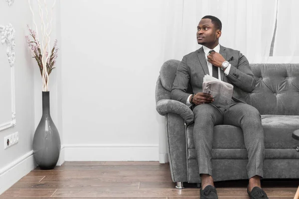 Young African American businessman in a gray suit reading a newspaper while sitting on a sofa.