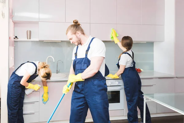 Servicio de limpieza con equipo profesional durante el trabajo. limpieza profesional de la cocina, limpieza en seco del sofá, lavado de ventanas y suelos. hombres y mujeres en uniforme, monos y guantes de goma — Foto de Stock