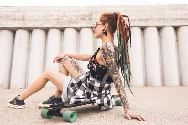 young girl with tattoo and dreadlocks sitting on a longboard on the background of concrete structure
