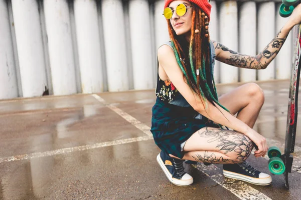 Young girl with tattoos and dreadlocks in a cap sits with a longboard on the background of concrete structure — Stock Photo, Image