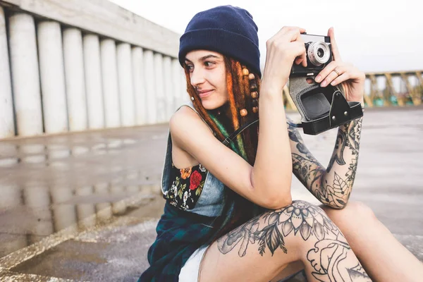 Chica joven con tatuajes y rastas en un sombrero azul fotografías de una cámara vintage en el fondo de una pared de hormigón —  Fotos de Stock